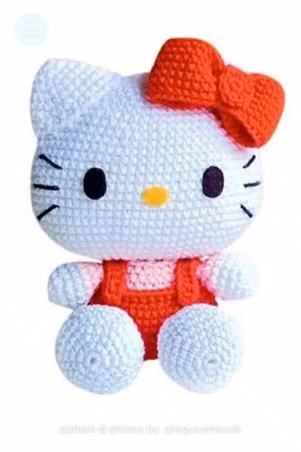 A crocheted Hello Kitty doll wearing red overalls and a red bow on her head, featured in a crochet roundup from https://theyarncrew.com/.