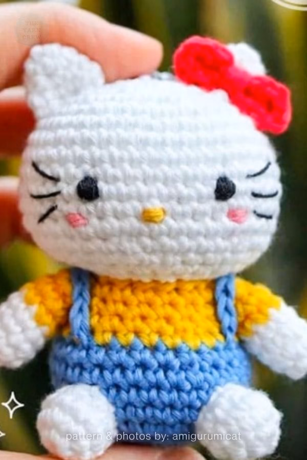 A close-up of a crocheted Hello Kitty doll held in a hand, wearing a yellow shirt and blue overalls with a bright red bow on her head, featured in a crochet roundup from https://theyarncrew.com/.