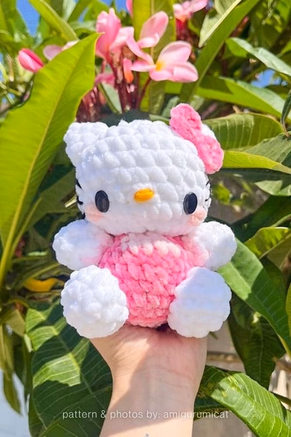 A crocheted Hello Kitty doll made of chunky plush yarn held in a hand outdoors against green leaves and pink flowers, wearing a light pink outfit and matching bow, featured in a crochet roundup from https://theyarncrew.com/.