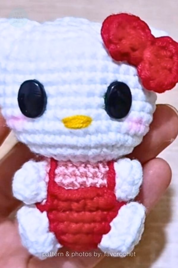 A close-up of a small crocheted Hello Kitty doll held in a hand, wearing a pink shirt and red overalls with a red bow on her head, featured in a crochet roundup from https://theyarncrew.com/.