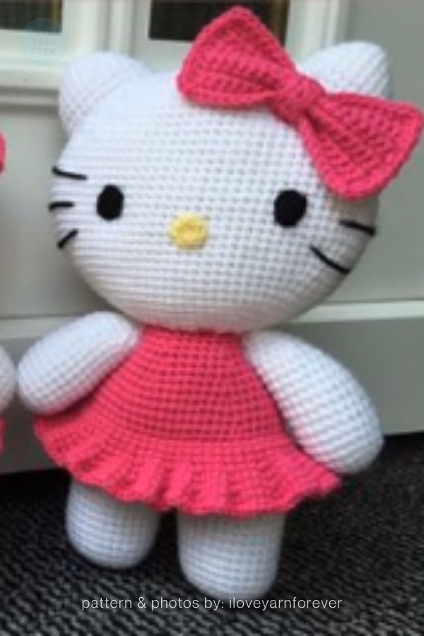 A crocheted Hello Kitty doll standing in a bright pink ruffled dress with a matching pink bow on her head, featured in a crochet roundup from https://theyarncrew.com/.