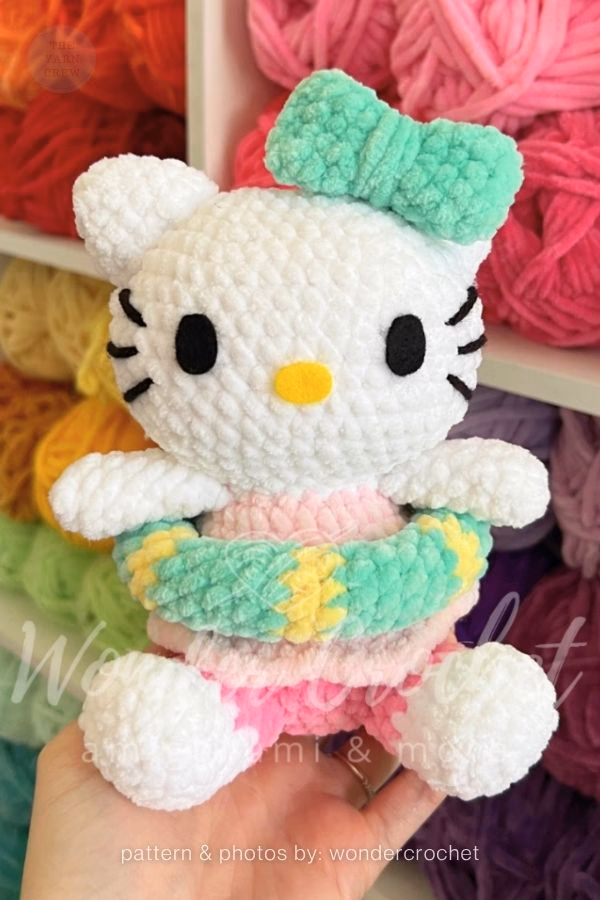 A crocheted Hello Kitty doll made of chunky plush yarn held in a hand, wearing a pink outfit, a green and yellow swim ring, and a green bow on her head, featured in a crochet roundup from https://theyarncrew.com/.