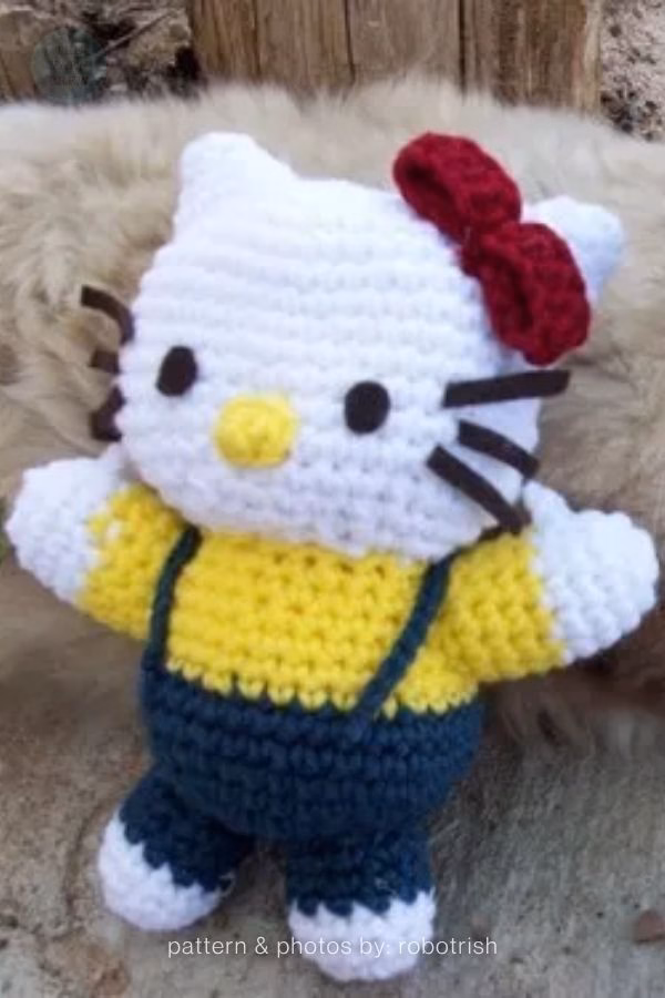 A crocheted Hello Kitty doll wearing a yellow shirt and dark blue overalls with a red bow on her head, featured in a crochet roundup from https://theyarncrew.com/.