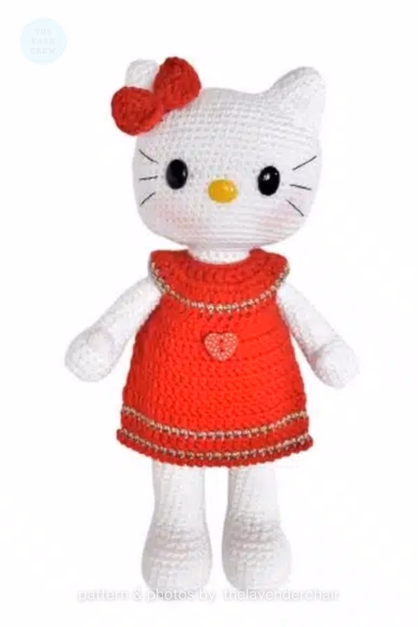 A crocheted Hello Kitty doll standing in a red dress with a heart button and a matching red head bow, featured in a crochet roundup from https://theyarncrew.com/.