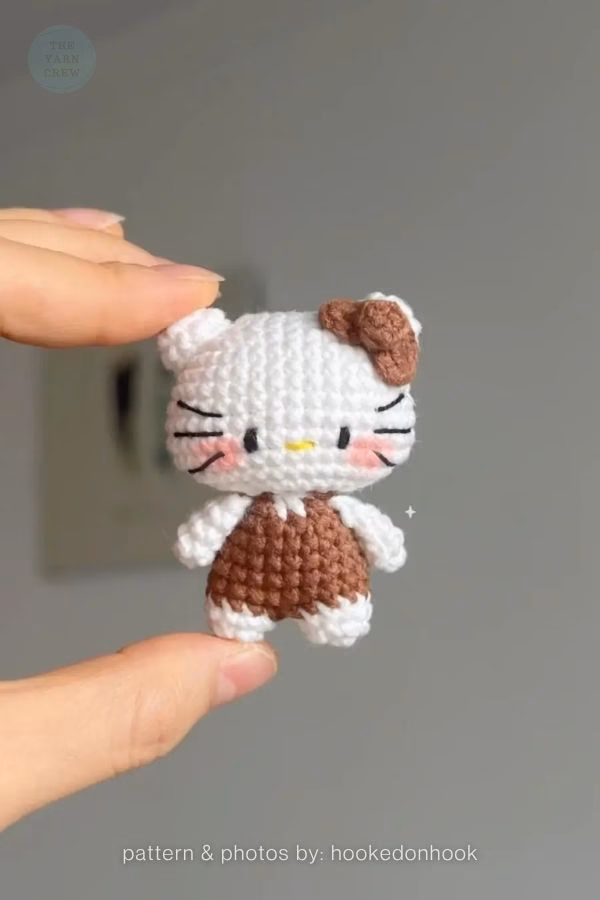 A tiny crocheted Hello Kitty doll held between two fingers, wearing a brown outfit and a matching brown bow, featured in a crochet roundup from https://theyarncrew.com/.