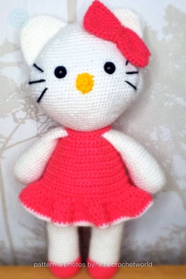 A crocheted Hello Kitty doll standing in a bright pink dress with a ruffled skirt and a matching pink bow on her head, featured in a crochet roundup from https://theyarncrew.com/.
