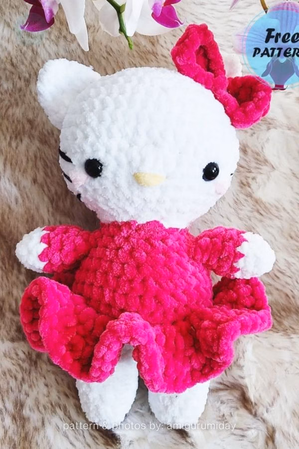 A crocheted Hello Kitty doll made of chunky plush yarn, wearing a bright pink ruffled dress and a matching bow, featured in a crochet roundup from https://theyarncrew.com/.