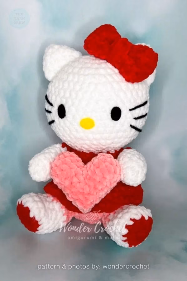 A crocheted Hello Kitty doll made of plush yarn sitting and holding a large pink heart, wearing a red outfit and matching red bow, featured in a crochet roundup from https://theyarncrew.com/.