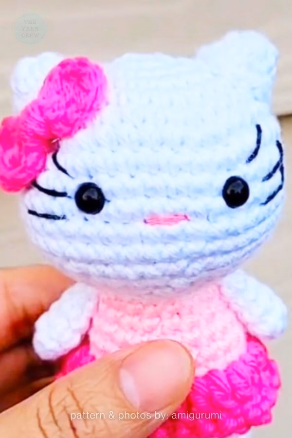 A small crocheted Hello Kitty doll held in a hand, wearing a light pink and bright pink ruffled dress with a matching pink bow, featured in a crochet roundup from https://theyarncrew.com/.