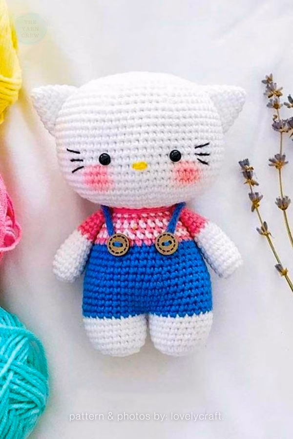 A crocheted Hello Kitty doll wearing a pink and white shirt under blue overalls with two small wooden buttons, featured in a crochet roundup from https://theyarncrew.com/.