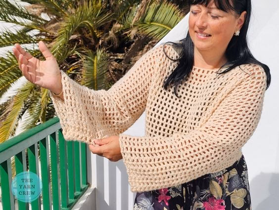 A woman standing outdoors near a green railing and a palm tree, modeling a beige, long-sleeved, cropped crochet mesh sweater and holding out one of the wide sleeves. She is wearing dark floral pants. The Yarn Crew logo is visible in the bottom left corner. This is part of a crochet roundup from https://theyarncrew.com/.