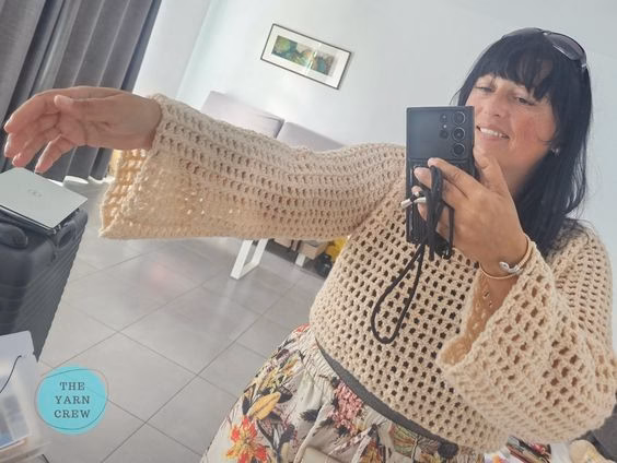 A woman taking a mirror selfie indoors, modeling a beige, long-sleeved, cropped crochet mesh sweater and floral print pants. She has one arm extended to the side to show off the wide sleeve. The Yarn Crew logo is visible in the bottom left corner. This is part of a crochet roundup from https://theyarncrew.com/.