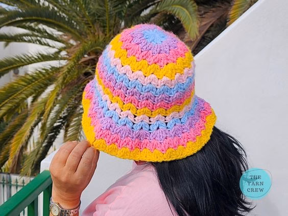 Colorful crochet bucket hat made with pink, yellow, blue, and purple yarn in a wavy stripe pattern, shown from the back. Original design created by Ariana Wimsett at TheYarnCrew.com.