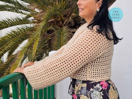 A woman standing outdoors in profile, holding onto a green railing near a palm tree. She is modeling a beige, long-sleeved, cropped crochet mesh sweater paired with dark floral print pants. The Yarn Crew logo is visible in the top right corner. This is part of a crochet roundup from https://theyarncrew.com/.