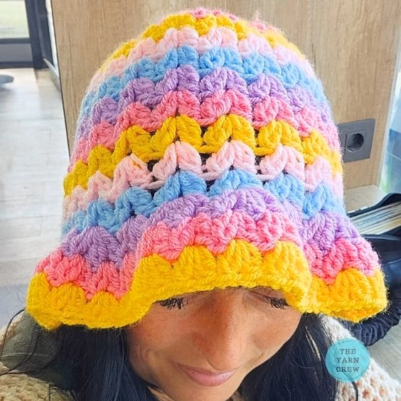 Colorful crochet bucket hat made with pink, yellow, blue, and purple yarn in a wavy stripe pattern, shown from a front angle looking down. Original design created by Ariana Wimsett at TheYarnCrew.com.