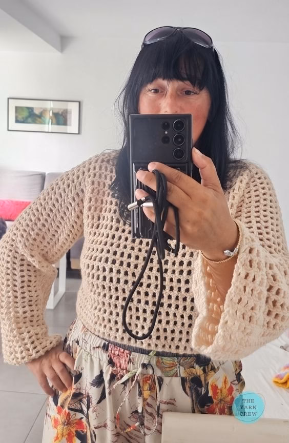 A woman taking a mirror selfie wearing a beige, long-sleeved, cropped crochet mesh sweater paired with floral print pants. The Yarn Crew logo is visible in the bottom right corner. This is part of a crochet roundup from https://theyarncrew.com/.