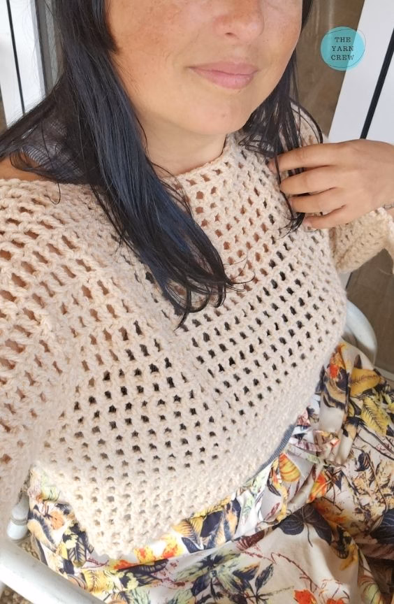 A close-up selfie of a woman wearing a beige, long-sleeved, cropped crochet mesh sweater paired with floral print bottoms. Her hand is resting near her shoulder, and The Yarn Crew logo is visible in the top right corner. This is part of a crochet roundup from https://theyarncrew.com/.