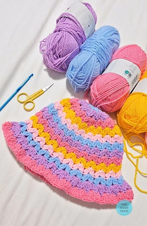Colorful crochet bucket hat in a wavy stripe pattern, displayed flat next to yarn skeins in pink, yellow, blue, and purple, a crochet hook, and scissors. Original design created by Ariana Wimsett at TheYarnCrew.com.