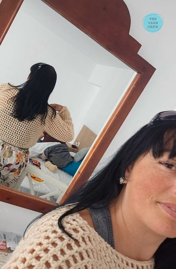 A woman taking a mirror selfie indoors, focusing on the back view of a beige, long-sleeved, cropped crochet mesh sweater that she is wearing. The reflection shows the back of the sweater and her floral pants, while her face and the shoulder of the sweater are visible in the foreground. The Yarn Crew logo is visible in the top right corner of the mirror. This is part of a crochet roundup from https://theyarncrew.com/.