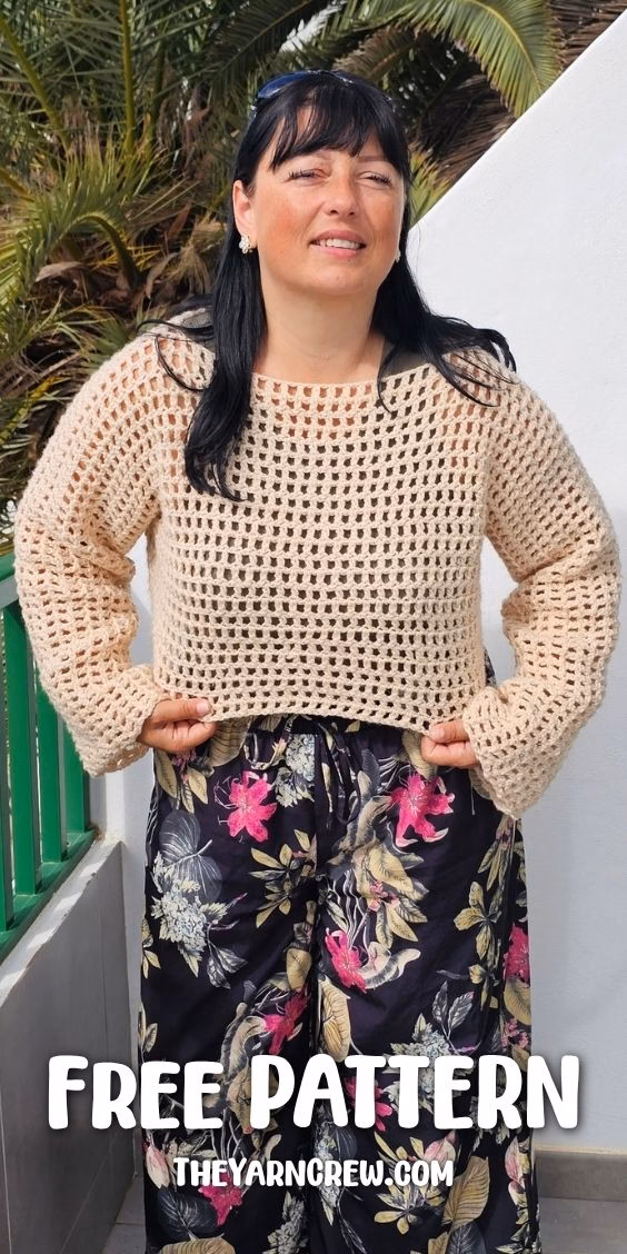 A woman standing outdoors near a green railing and palm tree, modeling a beige, long-sleeved, cropped crochet mesh sweater paired with dark floral print pants. She is holding the bottom hem of the sweater with both hands. The text on the image reads "FREE PATTERN" and "THEYARNCREW.COM". This is part of a crochet roundup from https://theyarncrew.com/.