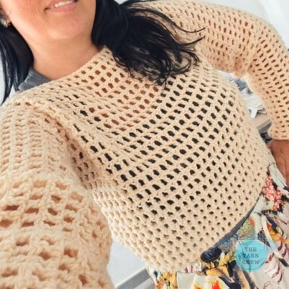 A close-up of a woman modeling a beige, long-sleeved, cropped crochet mesh sweater paired with floral pants. The Yarn Crew logo is visible in the bottom right corner. This is part of a crochet roundup from https://theyarncrew.com/.