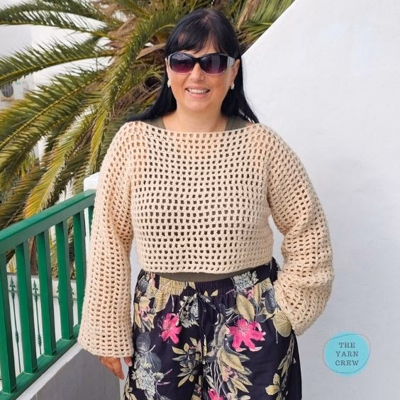 A woman standing outdoors near a green railing and a palm tree, smiling and wearing sunglasses. She is modeling a beige, long-sleeved, cropped crochet mesh sweater over an olive green top, paired with dark floral print pants. The Yarn Crew logo is visible in the bottom right corner. This is part of a crochet roundup from https://theyarncrew.com/.