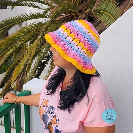 Colorful crochet bucket hat made with pink, yellow, blue, and purple yarn in a wavy stripe pattern, shown from a side profile view. Original design created by Ariana Wimsett at TheYarnCrew.com.