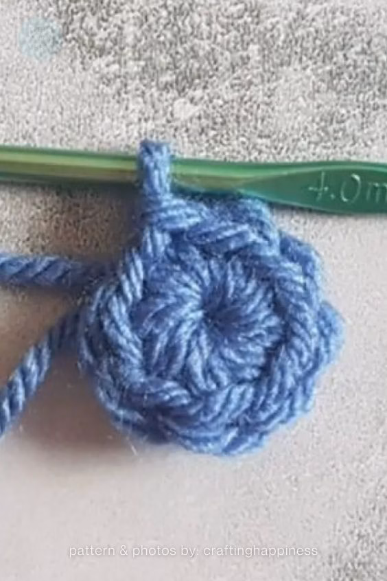 Close-up shot of a completed blue yarn crochet magic ring on a gray background, with a green 4.0mm crochet hook resting at the top. Original Design by Ariana of CraftingHappiness.