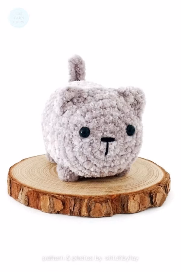 A small, round amigurumi cat made from soft, light gray velvet yarn sitting on a natural wood slice; from a roundup on TheYarnCrew by Ariana Wimsett.