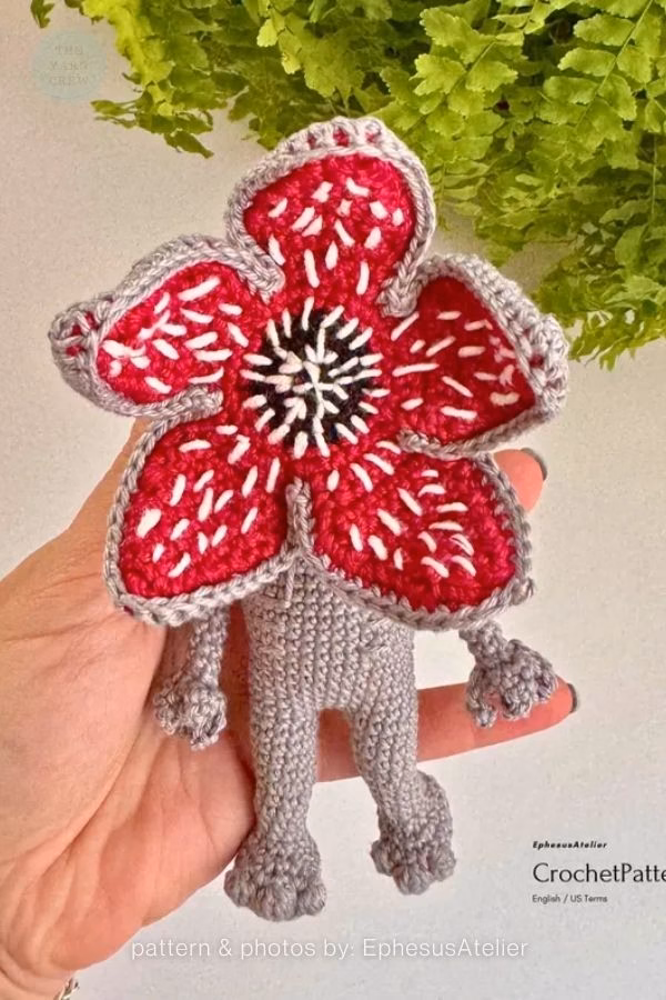 A hand holding a grey crochet Demogorgon toy from Stranger Things with its giant, red flower-like face open. Curated by Ariana of TheYarnCrew.