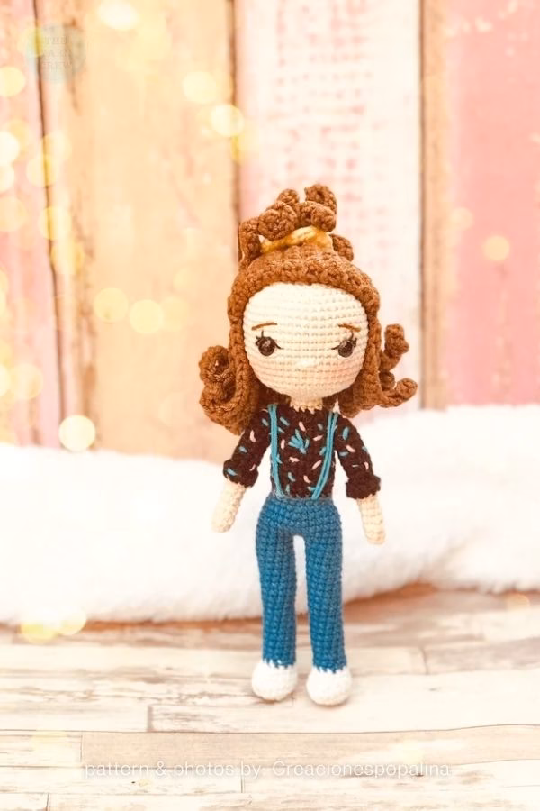 A slender crochet doll of Eleven from Stranger Things with curly brown hair tied in a scrunchie, wearing a patterned shirt and blue jeans with suspenders. Curated by Ariana of TheYarnCrew.