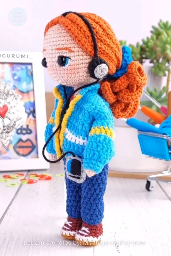 A side view of a crochet doll of Max from Stranger Things with thick orange hair, a blue jacket, headphones, and a tiny cassette player. Curated by Ariana of TheYarnCrew.