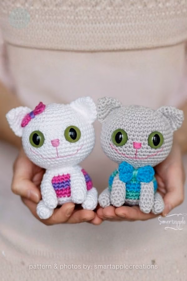 A pair of small amigurumi cats, one white with a pink bow and one gray with a blue bowtie, held in two hands; from a roundup on TheYarnCrew by Ariana Wimsett.