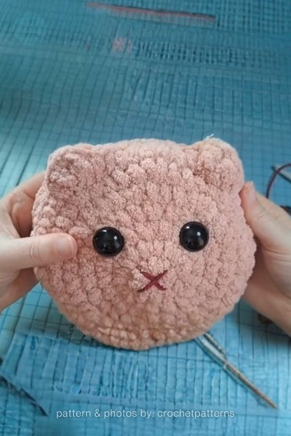 A close-up of a round, pink cat head crocheted with thick, textured velvet yarn featuring large black safety eyes; from a roundup on TheYarnCrew by Ariana Wimsett.