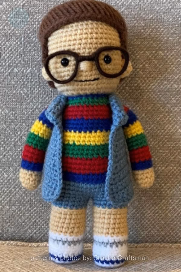 A crochet doll of Derek from Stranger Things wearing a colorful rainbow-striped shirt, blue vest, and round glasses. Curated by Ariana of TheYarnCrew.