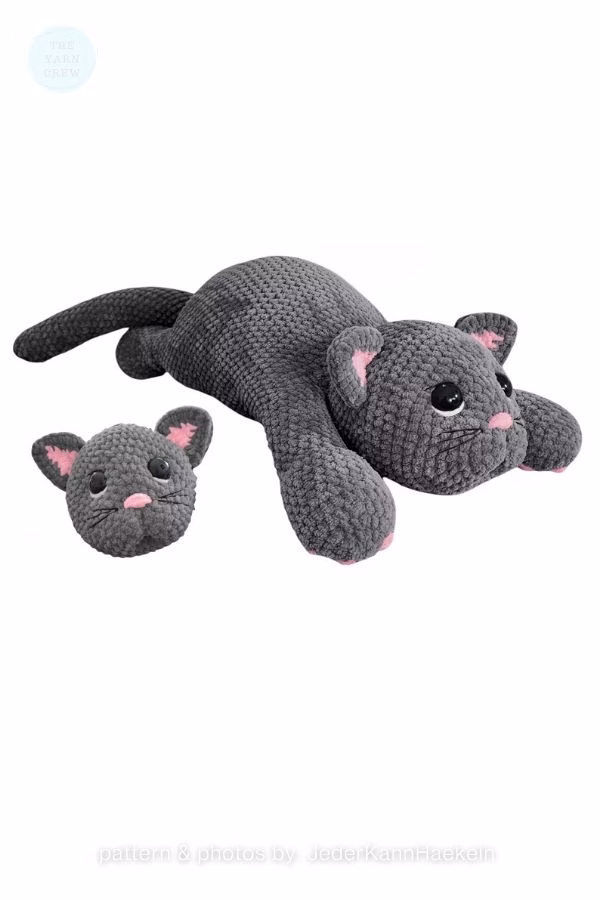A large, floppy dark gray crochet cat lying flat next to a smaller matching cat head, both featuring pink ears and noses; from a roundup on TheYarnCrew by Ariana Wimsett.