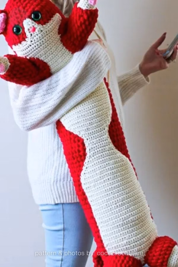 A person holding a very long, oversized red and white crochet cat designed to be a body pillow; from a roundup on TheYarnCrew by Ariana Wimsett.