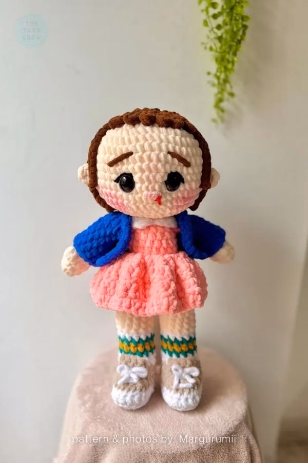 A soft crochet doll of Eleven from Stranger Things wearing her classic pink dress, blue jacket, and sneakers. Curated by Ariana of TheYarnCrew.