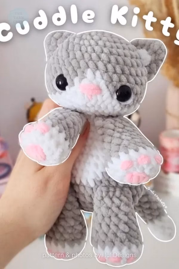 A handmade "Cuddle Kitty" amigurumi crocheted with chunky gray and white yarn and pink paw pads; from a roundup on TheYarnCrew by Ariana Wimsett.