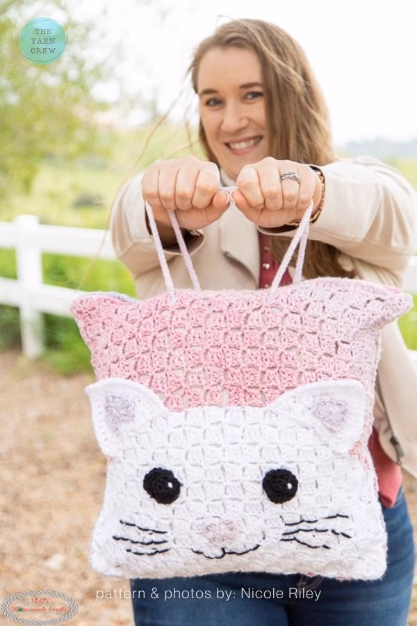 A woman holding a large, square crochet tote bag featuring a white cat face on a pink ombre background; from a roundup on TheYarnCrew by Ariana Wimsett.
