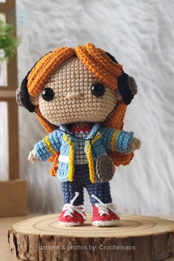 A cute crochet doll of Max from Stranger Things with bright orange hair, a blue jacket, and black headphones. Curated by Ariana of TheYarnCrew.