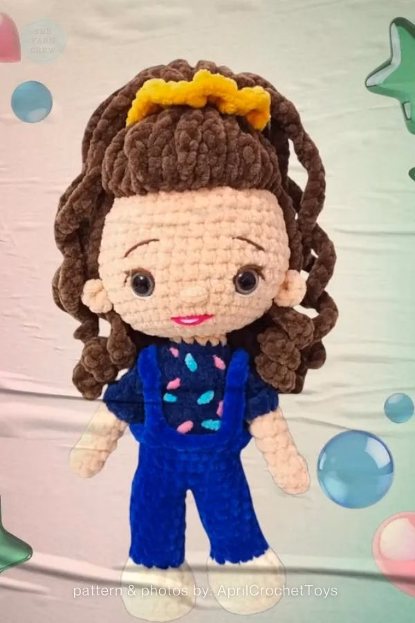 A fluffy crochet doll of Eleven from Stranger Things with curly brown hair, a yellow scrunchie, and blue overalls. Curated by Ariana of TheYarnCrew.