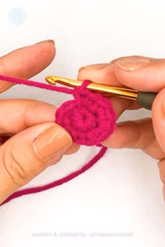 Close-up of a person's hands using a gold crochet hook to work bright pink yarn into a small crochet magic ring. Curated by Ariana of TheYarnCrew.