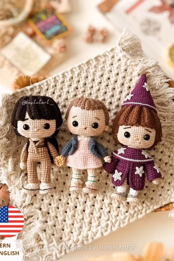 Three small crochet dolls from Stranger Things holding hands, featuring Mike in a tan jumpsuit, Eleven in a pink dress, and Will in a purple wizard costume. Curated by Ariana of TheYarnCrew.
