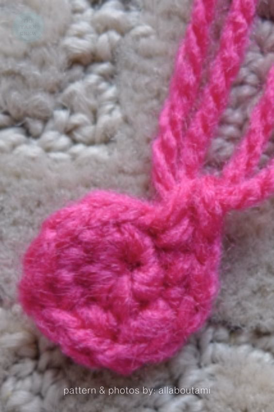 Close-up shot of a small, bright pink crocheted circle with long yarn tails resting on a textured, neutral-colored carpet. Curated by Ariana of TheYarnCrew.