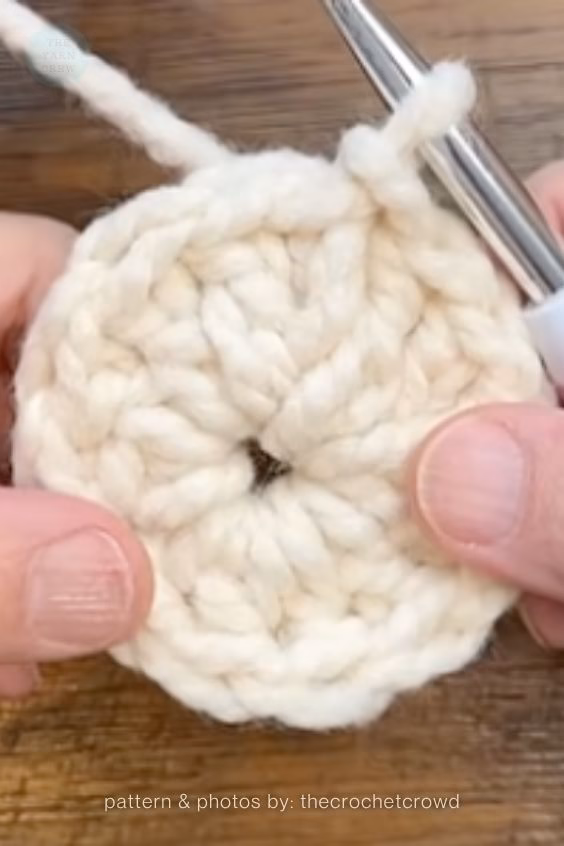 Close-up of hands holding a thick, cream-colored crocheted magic ring on a wooden surface, with a silver crochet hook inserted into the top stitch. Curated by Ariana of TheYarnCrew.