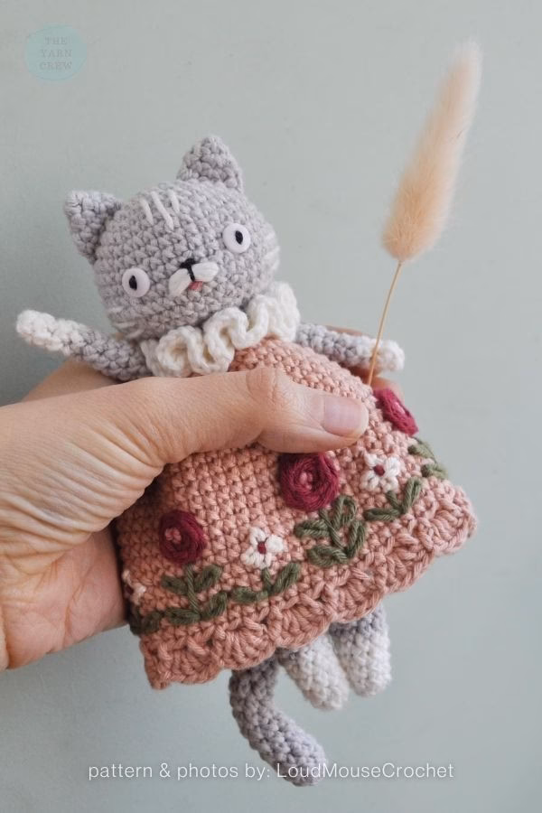 A whimsical crochet cat doll wearing a dusty pink dress with embroidered red flowers; from a roundup on TheYarnCrew by Ariana Wimsett.