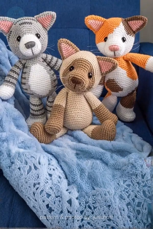 Three realistic amigurumi cats—a gray tabby, a Siamese, and an orange tabby—sitting on a blue blanket; from a roundup on TheYarnCrew by Ariana Wimsett.