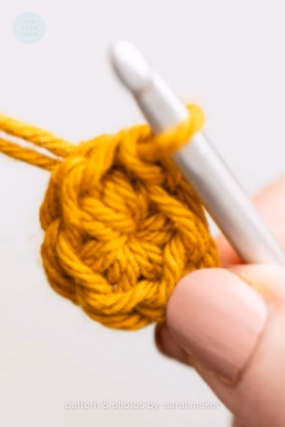 Extreme close-up of fingers with a soft pink manicure holding a small, tightly closed crochet magic circle made from mustard yellow yarn, with a silver hook visible. Curated by Ariana of TheYarnCrew.