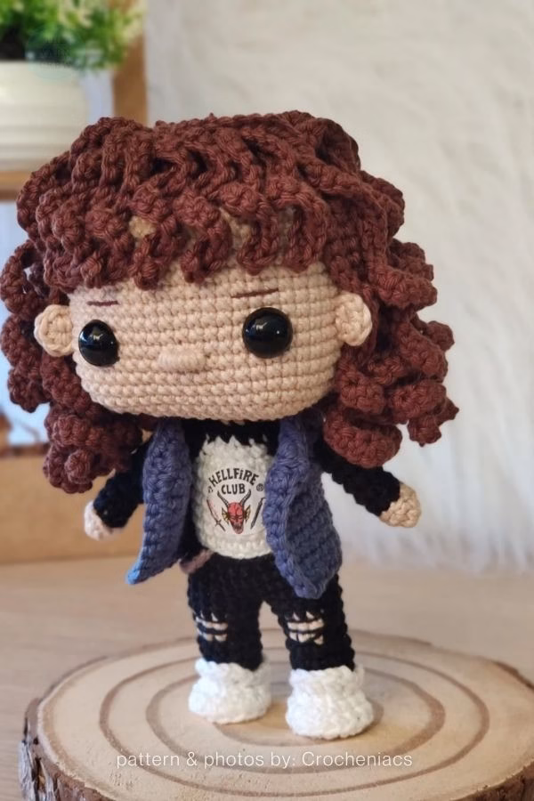 A crochet doll of Eddie Munson from Stranger Things with curly brown hair, a denim vest, and a tiny Hellfire Club t-shirt. Curated by Ariana of TheYarnCrew.